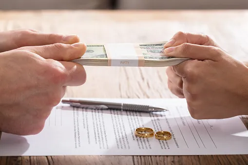 Stack of money being pulled by hand on opposite sides over divorce papers and wedding bands signifying arguing over money in a divorce.