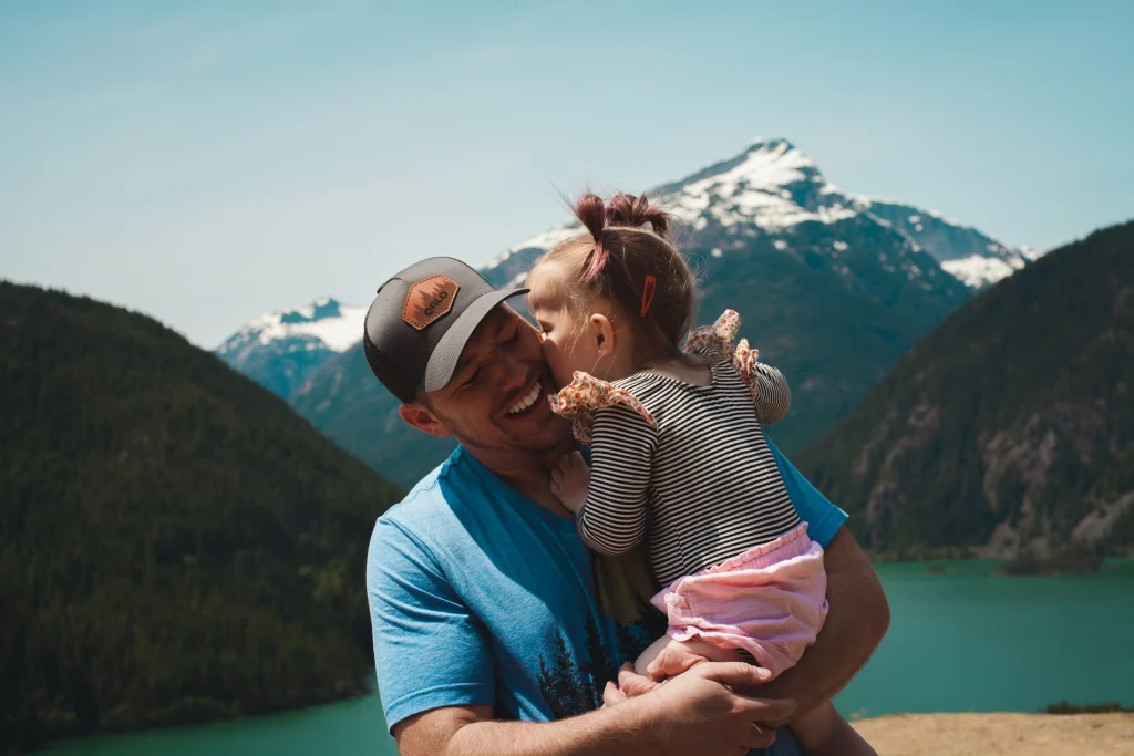 Father holds child in front of a mountain and lake as they laugh. 