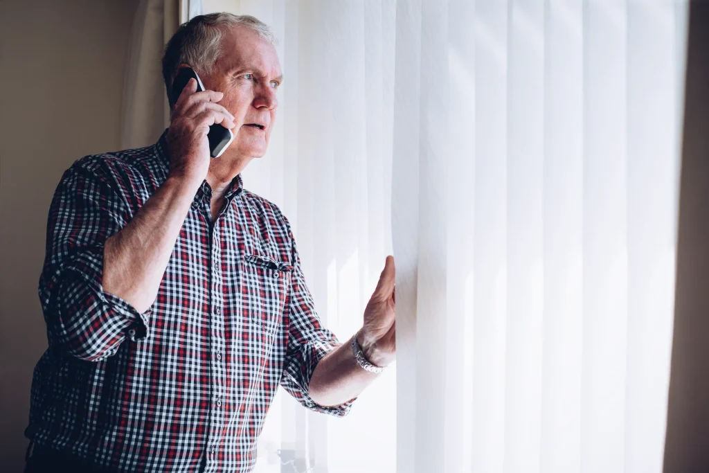 Man peering through vertical blinds while talking on the phone.